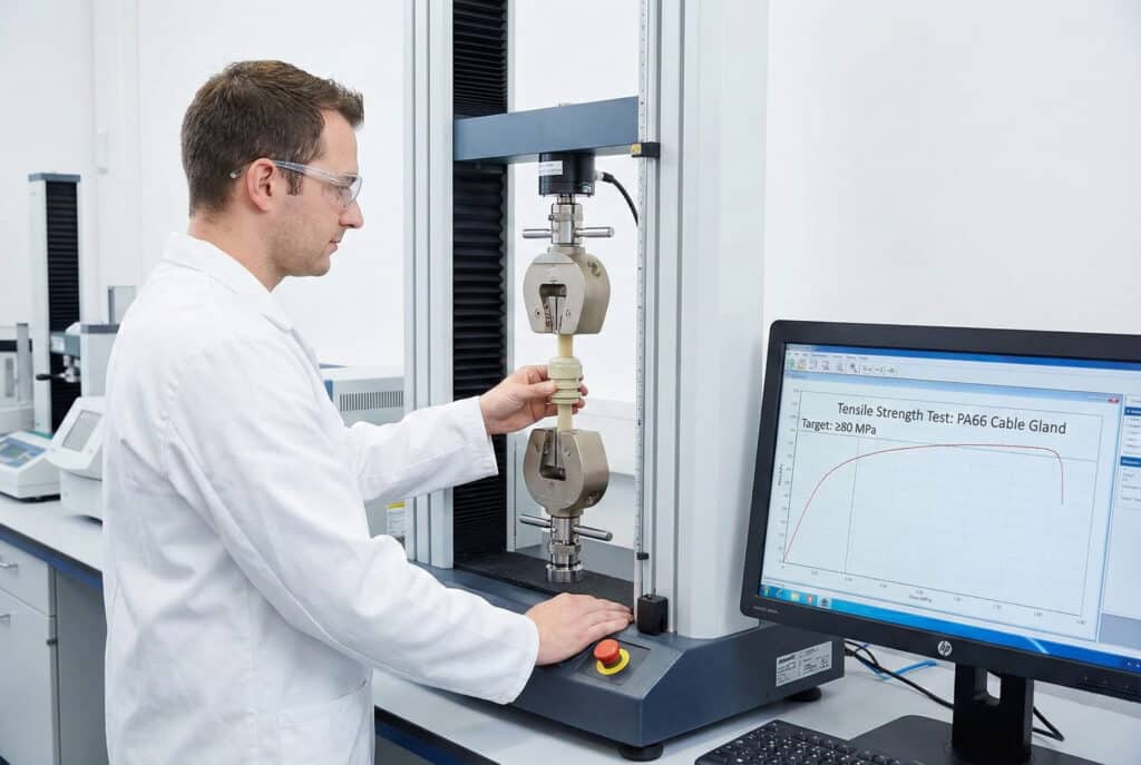 A laboratory technician in a white coat and safety glasses performs a tensile strength test on a PA66 cable gland using a materials testing machine. A computer monitor next to the machine displays a graph with the text "Tensile Strength Test: PA66 Cable Gland" and "Target: ≥80 MPa."