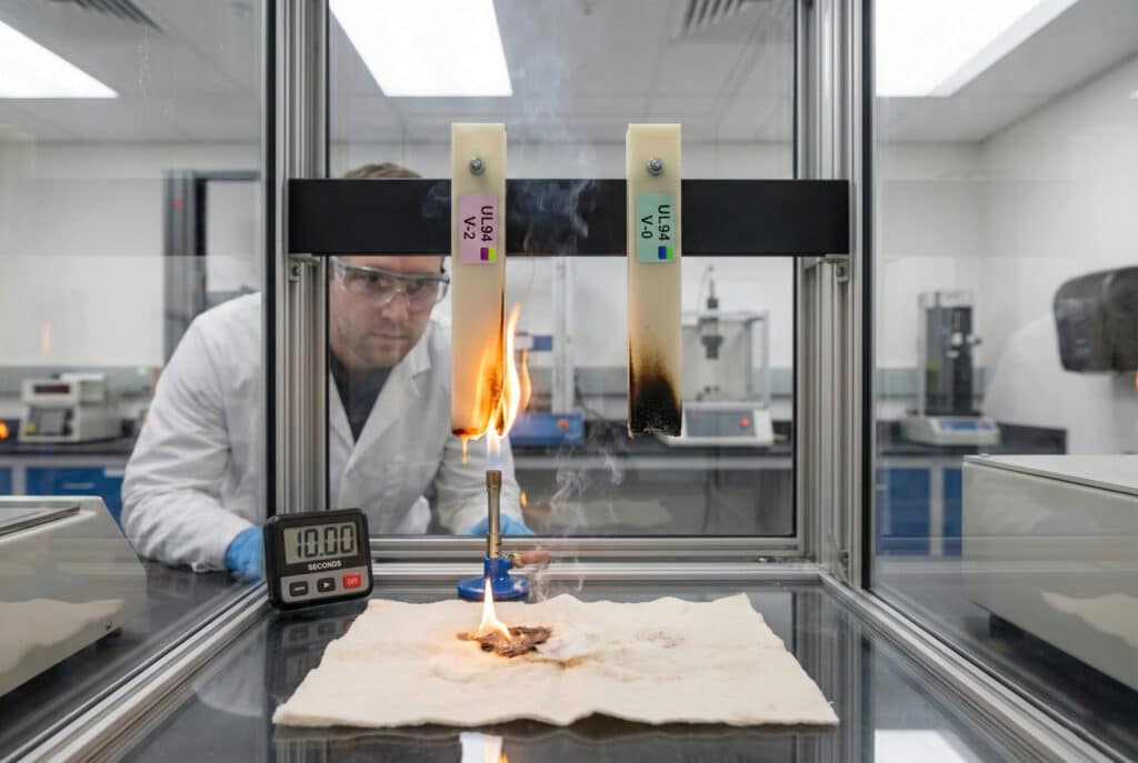 Technik v laboratórnej digestore vykonáva vertikálny test horľavosti UL94 na dvoch vzorkách nylonu označených ako 'UL94 V-2' a 'UL94 V-0'. Vzorka V-2 naľavo horí a kvapky ohňa zapália bavlnenú podložku pod ňou, zatiaľ čo vzorka V-0 napravo sa sama uhasila a má ohorený okraj. Časomer ukazuje 10 sekúnd.